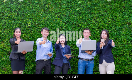 Team of Asian business man and woman holding tablet and celebrating success of business plan outside office at green leaf wall Stock Photo