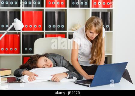 Tired young business woman is sleeping on workplace in the office, another employee took advantage of the situation and is stealing private information from computer of competitor. Stock Photo