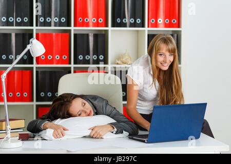 Tired young business woman is sleeping on workplace in the office, another employee took advantage of the situation and is stealing private information from computer of competitor. Stock Photo