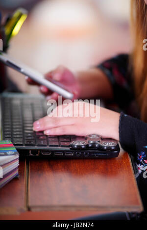 Young woman using her cellphone in the office and a popular fidget spinner toy over the laptop, on office background. Stock Photo