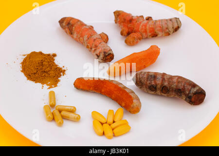Turmeric ground into powder, in capsule and pill form, and the whole raw root or rhizome peeled, unpeeled, partially peeled, on a round white plate with an orange background. Stock Photo