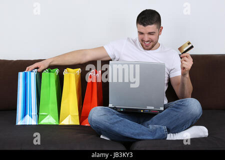 Ein junger Mann sitzt auf dem Sofa mit Einkaufstüten und kauft im Internet ein Stock Photo