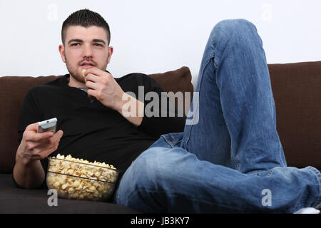 Ein junger Mann sitzt auf dem Sofa, schaut fernsehen und isst Popcorn Stock Photo