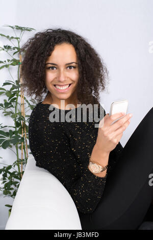 Junge, hübsche Frau sitzt auf dem Sofa und verwendet ein Smartphone Stock Photo