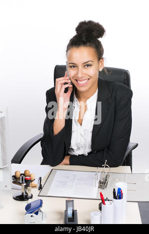 Junge, hübsche Büroangestellte sitzt am Schreibtisch im Büro und telefoniert mit einem Smartphone Stock Photo