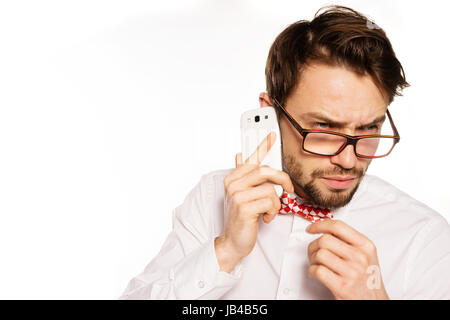 Nerdy bearded businessman wearing glasses and a polka dot bow tie frowning as he listens to the conversation while talking on a smartphone, isolated on white Stock Photo