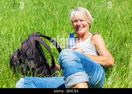 smiling mature woman with a bottle in the hand lying in the meadow at a hiking rest Stock Photo