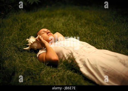 Young innocent woman lying on grass listening to seashell. 16, 17, 18, 19, 20, 21, 25, years, years old Stock Photo