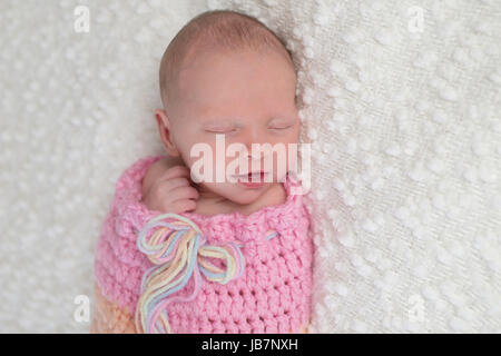 Headshot of a smiling three week old newborn baby girl bundled up in a light pink, crocheted, snuggle sack. She is lying on a white, bouncle blanket. Stock Photo