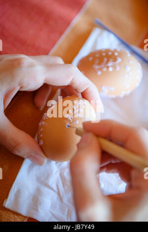 Traditional decorating of the easter eggs by the woman, visible woman's hands, in the background visible another easter egg, shallow DOF. Stock Photo