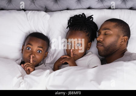 High angle portrait of siblings with finger on lips by father sleeping in bed at home Stock Photo