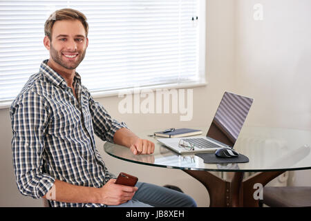 Handsome young white man sitting indoors in his house at his glass desk behind his modern notebook and technology, with his body turned sideways towar Stock Photo