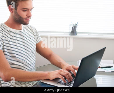 Attractive young caucasian male student wearing a black and white striped t-shirt, working on his modern laptop in the bright living room next to the  Stock Photo