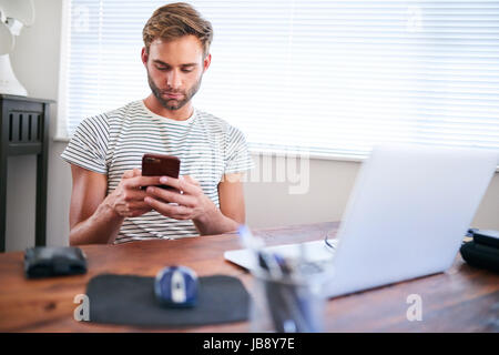 Good looking young caucasian guy busy using his phone with both hands while sitting at home behind his computer at his desk next to a nice large windo Stock Photo