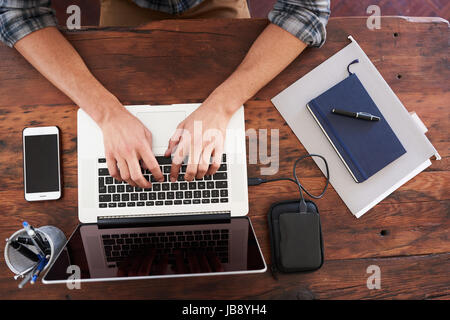 Birds eye view of hands typing on a modern notebook, placed on a durable dark wood desk along with some notes, portable storage, a pen holder and a mo Stock Photo