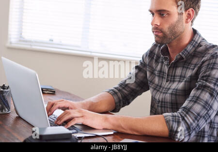 Young adult man looking frustrated and fed-up with work, while still typing on his notebook, seated at his wooden desk with a large bright window maki Stock Photo