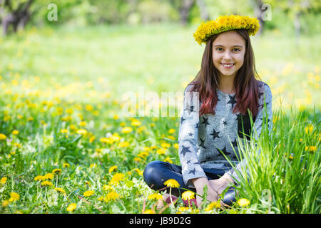 Cute young girl wearing wreath of dandelions and smiling while sitting on grass in park Stock Photo