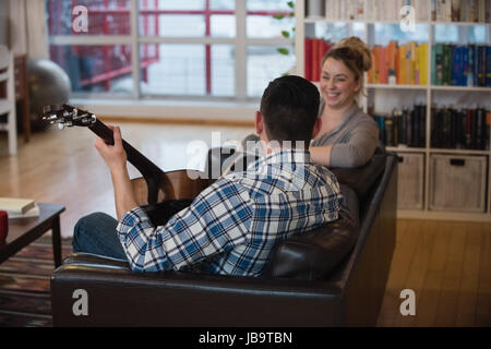 Man playing guitar while woman sitting beside him in living room at home Stock Photo