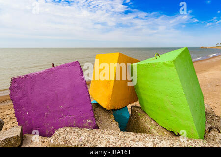 Three different colours - purple, yellow and green concrete breakwater blocks on the beach, in the backgroundblue cloudless sky, yellow sand and coastline. Stock Photo