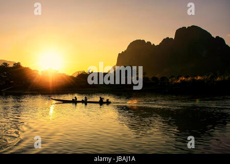 Sunset over Nam Song River with silhouetted rock formations and a boat in Vang Vieng, Laos. Vang Vieng is a popular destination for adventure tourism  Stock Photo