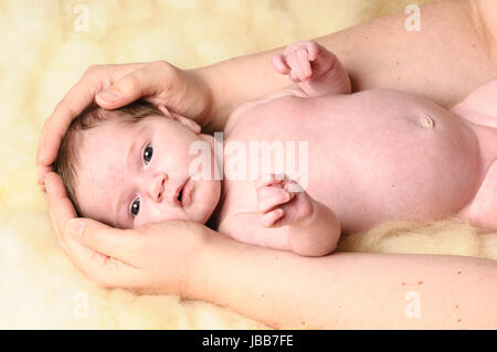 Ein kleines neugeborenes Baby liegt auf einem Schaffell. Sie Haende seiner Mutter umfassen zaertlich seinen Kopf.