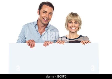 Lovely couple standing behind blank billboard, smiling and looking at the camera. Stock Photo