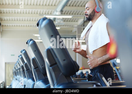Side view portrait of muscular sportsman running on treadmill listening to music during intense cardio workout in modern gym Stock Photo