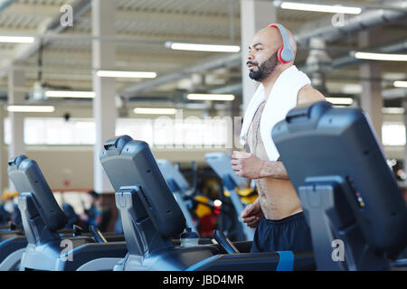 Side view portrait of muscular sportsman running on treadmill listening to music during cardio workout in modern gym Stock Photo
