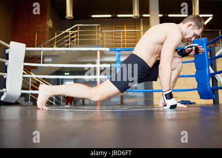 Portrait of muscular athlete warming up before sports training, stretching legs on floor in boxing ring