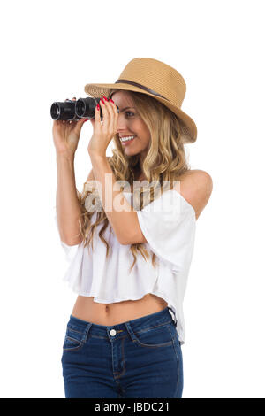 Smiling blond young woman in straw hat, jeans and white shirt looking away through binoculars. Waist up studio shot on isolated on white. Stock Photo