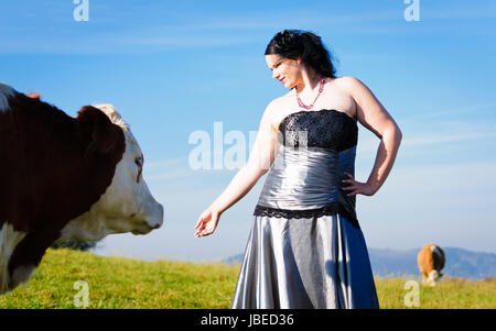 Erwachsene Frau im eleganten Abendkleid steht mitten auf einer Vieweide und streichelt liebevoll die Kühe. Stock Photo