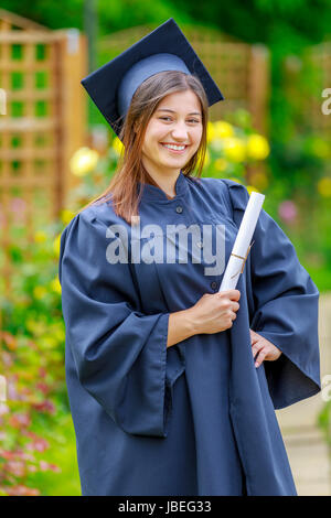 Smiling young woman holding diploma and wearing cap and gown outdoors looking at camera. Graduation concept. Stock Photo