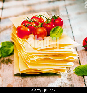 Dried uncooked lasagne pasta sheets with fresh ingredients including cherry tomatoes and basil leaves, close up on old wooden boards Stock Photo