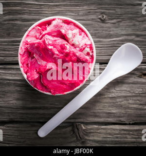 Delicious cold refreshing berry ice cream in a takeaway plastic tub with a teaspoon on an old textured wooden surface, overhead view in square format Stock Photo