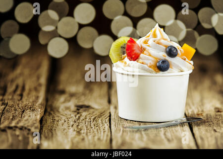 Creamy vanilla frozen yogurt topped with fresh tropical fruit served in a plastic takeaway tub with a disposable teaspoon on a wooden table with a background bokeh of party lights Stock Photo