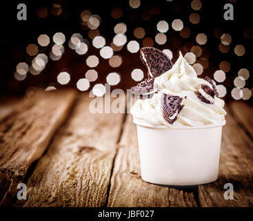 Delicious party dessert with frozen yogurt and chocolate oreos in a plastic takeaway tub standing on an old wooden counter with a twinkling bokeh of festive lights Stock Photo