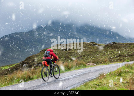 Lonely biker cycling through the Scottish Highlands on heavily laden touring bicycle along single track road during sleet in spring, Scotland, UK Stock Photo