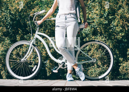 Slender young woman in a white T-shirt and jeans stands near her stylish bicycle in the summer in the park Stock Photo