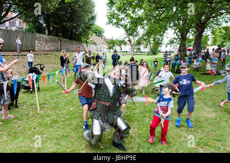 Medieval living history re-enactment event at Sandwich town, England. Man in knight's armour being cut down from behind by children with play swords. Small roped off arena, people watching. Stock Photo