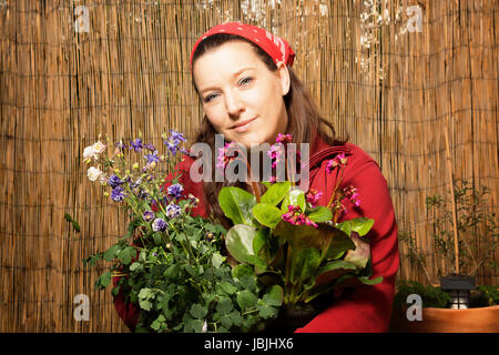 Woman with headscarf gardening and holding flower pots with different plants in front of a bamboo fence. She is happy about the beautiful plants. Stock Photo