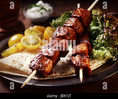 Healthy barbecued lean cubed pork kebabs served with a corn tortilla and fresh lettuce and tomato salad, close up view on a dark background Stock Photo