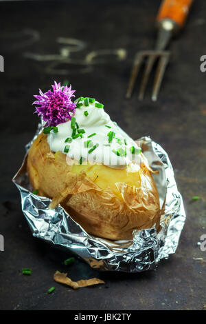 Foil baked jacket potato served in the aluminium foil wrapper topped with sour cream and fresh chopped chives and garnished with a purple chive flower Stock Photo