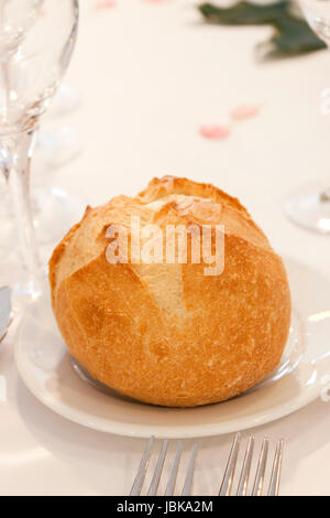 A bread bun on a plate on an elegant table with some cutlery and glass over a white cloth. Stock Photo