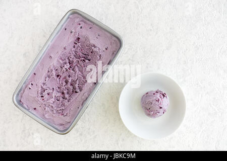 Homemade Blueberry Ice Cream in metal container and a big scoop in a white porcelain bowl on white marbled background with copy space. Stock Photo