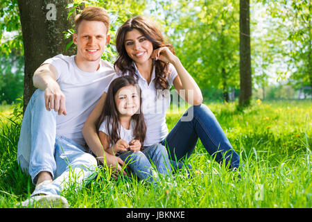 Portrait of happy family sitting on grass under a tree in park on a sunny day Stock Photo