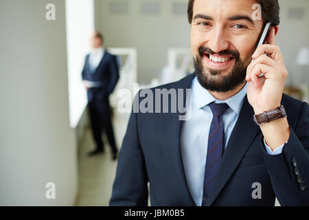 Image of young businessman speaking on the phone and looking at camera on background of his colleague Stock Photo
