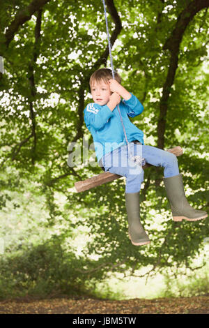 Boy in wellies swinging on tree rope swing Stock Photo