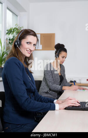Eine junge Büroangestellte mit Headset tippt auf einem Notebook. Eine weitere Frau mit Headset befindet sich im Hintergrund (unscharf). Stock Photo
