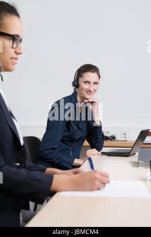 Eine junge Büroangestellte mit Headset sitzt vor einem Notebook im Büro. Eine weitere Frau befindet sich im Vordergrund (unscharf) und schreibt in ein Dokument. Stock Photo