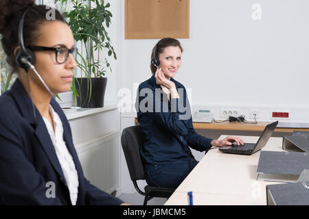 Eine junge Büroangestellte mit Headset sitzt vor einem Notebook im Büro. Eine weitere Frau mit Headset befindet sich im Vordergrund (unscharf). Stock Photo